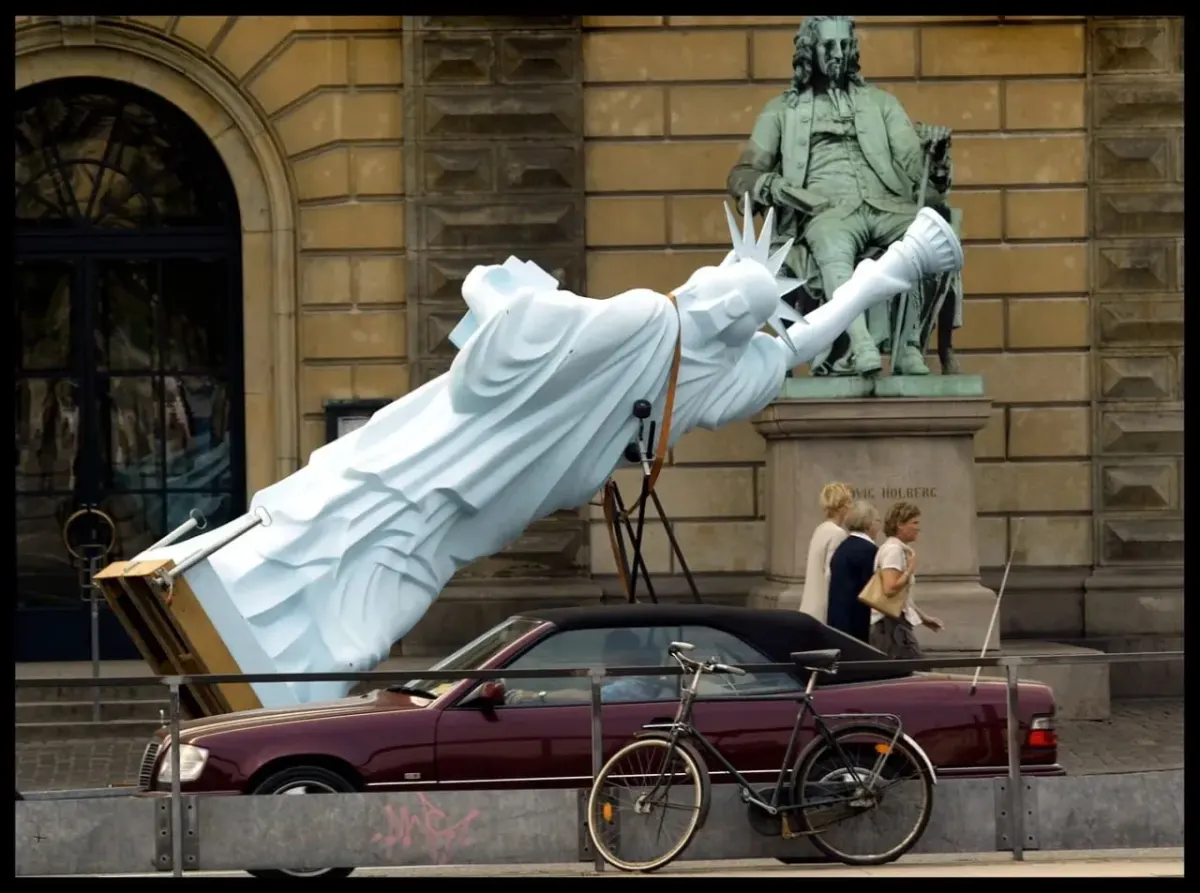 Street scene with a toppled Statue of Liberty sculpture near a building and a parked convertible and bicycle, suggesting complex cross-border issues